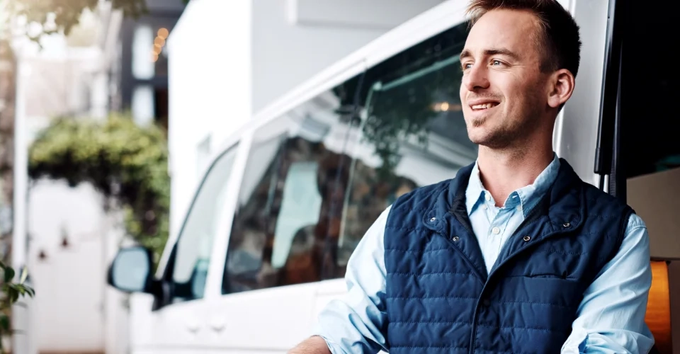 Homem sorridente junto a uma carrinha branca, simbolizando um serviço de transporte ou motorista profissional.