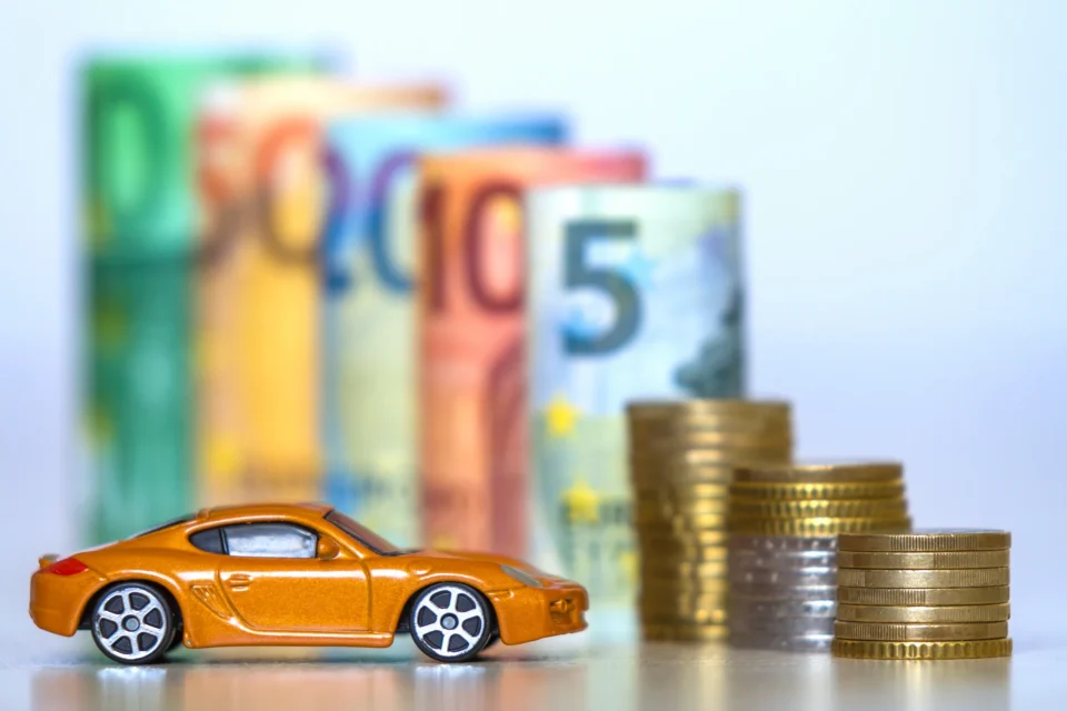 Carrinho em miniatura ao lado de pilhas de moedas e notas de euro desfocadas ao fundo, simbolizando o custo de um carro.